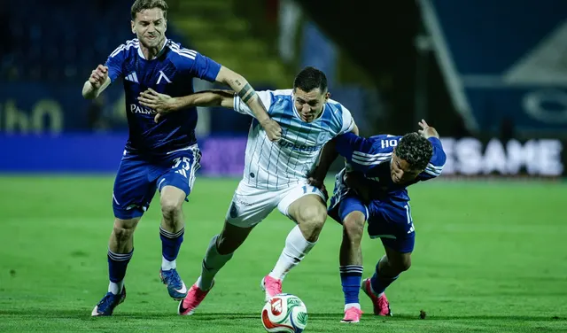 Fotbalist în echipament alb, între doi adversari în albastru închis, luptând pentru minge pe gazon