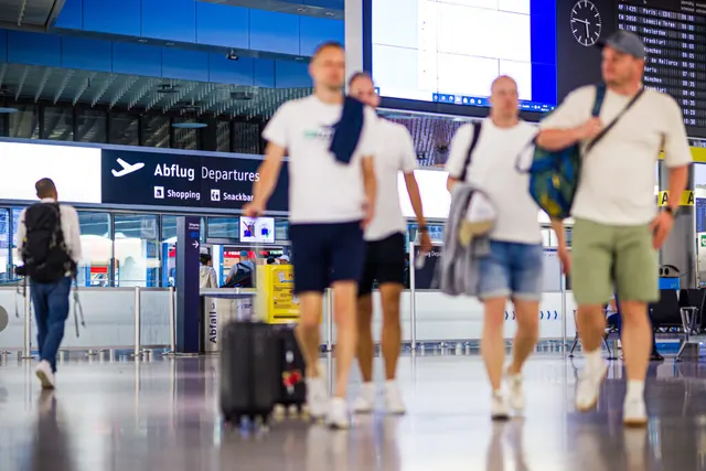 Călători cu bagaje într-un terminal de aeroport, sub panoul „Abflug Departures”