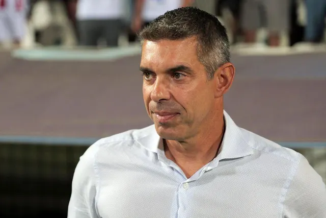 Bărbat cu părul grizonat, în cămașă albă, privind în lateral pe un stadion