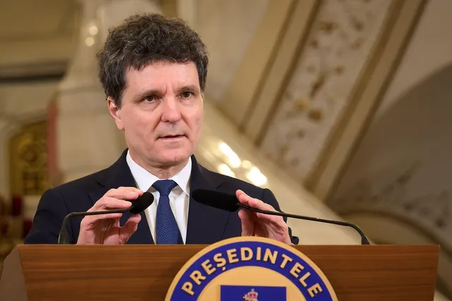 Bărbat cu părul ondulat, în costum, vorbind la pupitrul cu stema Președintelui României