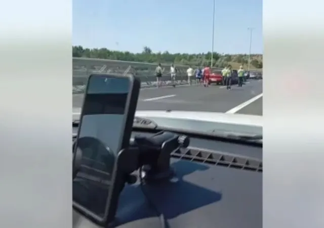 Telefon fixat pe bordul unei mașini; în față, mașini oprite pe autostradă și oameni pe carosabil