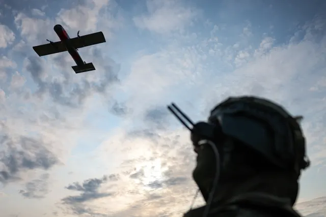 Dronă cu aripă fixă pe cer, urmărită de un militar cu cască, văzut din spate, în contralumină