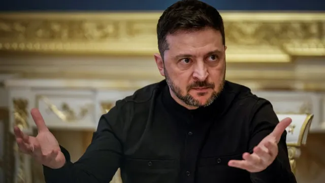 Bărbat cu barbă, în cămașă neagră, gesticulând cu ambele palme deschise, într-o sală ornată