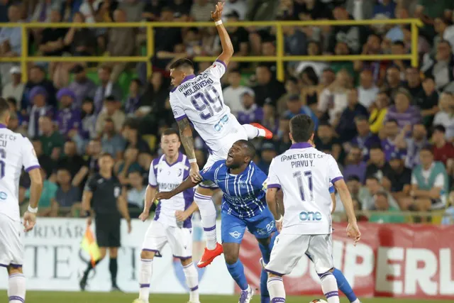 Fotbalist în echipament alb sărind la cap, printre jucători în alb și albastru, cu tribuna plină în spate