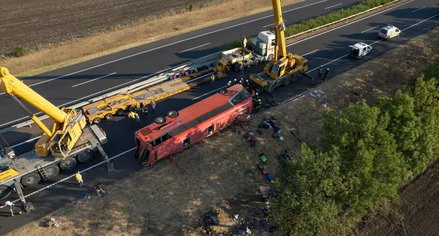 Autocar roșu răsturnat lângă o autostradă, ridicat de macarale galbene, văzut de sus