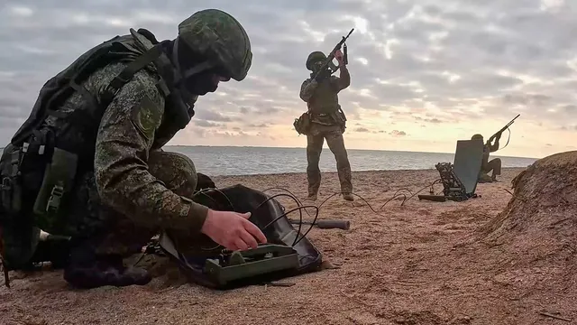 Militari pe o plajă la apus: unul îngenuncheat lângă un echipament electronic, altul cu arma ridicată