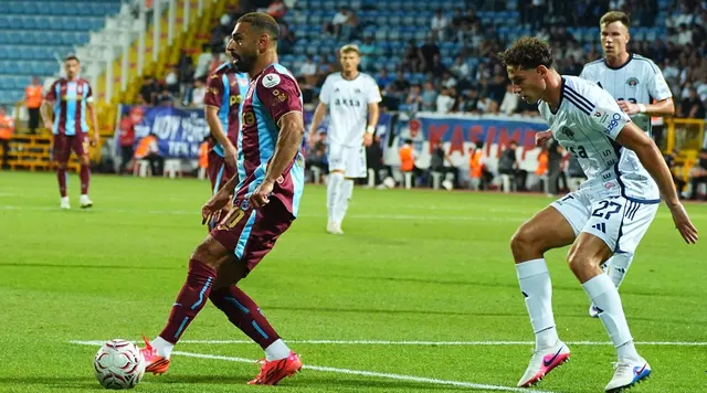Fotbalist în echipament vișiniu-albastru conducând mingea, marcat de un adversar în alb cu numărul 22