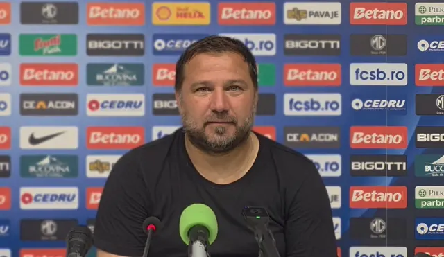 Bărbat cu barbă, în tricou negru, la o conferință de presă în fața panoului cu sponsorii FCSB