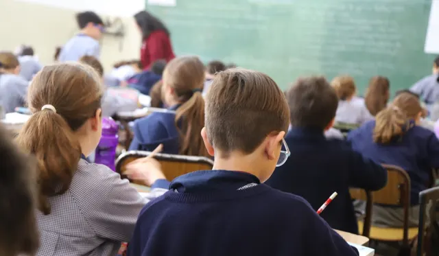 Elevi în bănci, văzuți din spate, într-o clasă, cu profesoara la tablă