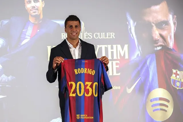 Bărbat în costum prezentând un tricou al Barcelonei cu inscripția „Rodrigo 2030”