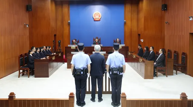 Bărbat în picioare între doi polițiști, în fața completului de judecată, într-o sală de tribunal chineză
