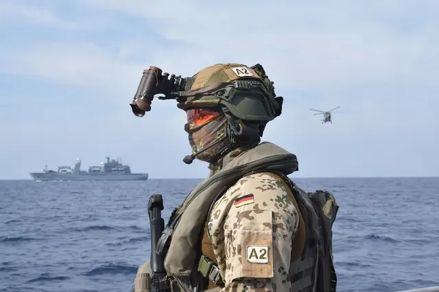 Militar cu cască și echipament de luptă pe mare, cu o navă și un elicopter în fundal