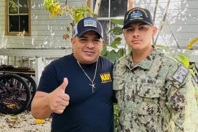 Bărbat cu șapcă ridicând degetul mare, lângă un marinar în uniformă de camuflaj a U.S. Navy