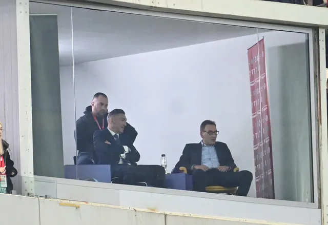Trei bărbați într-o cabină cu geam, în tribuna unui stadion