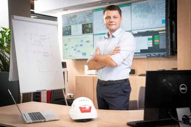Bărbat în cămașă albă, cu brațele încrucișate, într-o cameră de control cu monitoare și o cască albă pe birou