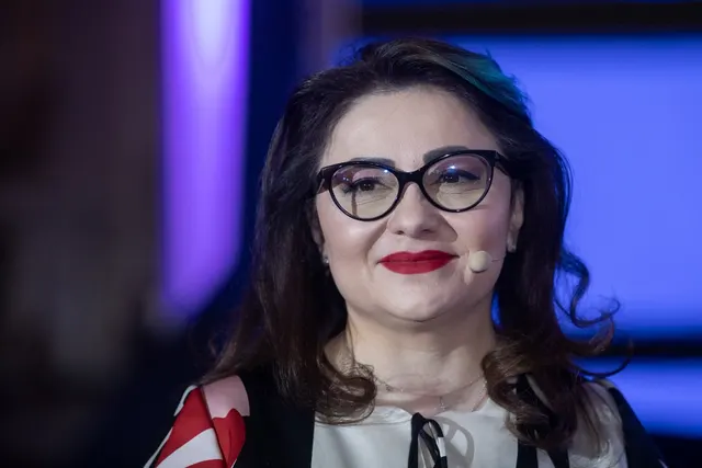Femeie cu ochelari și păr negru, cu microfon headset, zâmbind pe o scenă luminată în albastru