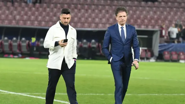 Doi bărbați traversând gazonul unui stadion: unul în sacou alb, cu telefonul, altul în costum albastru