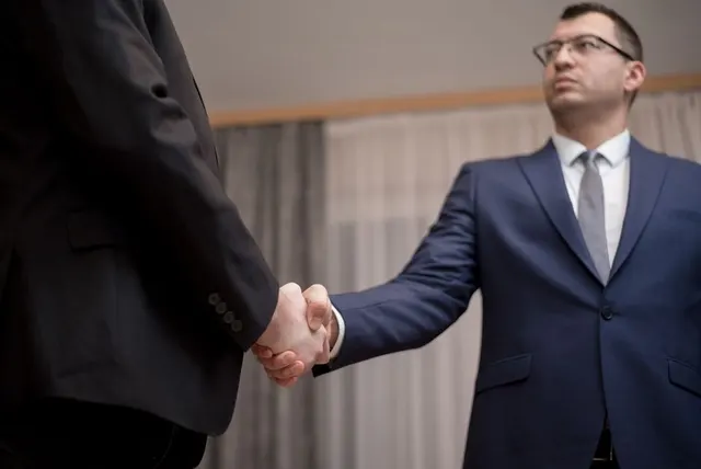Strângere de mână între doi bărbați în costume; unul, cu ochelari, privește în sus