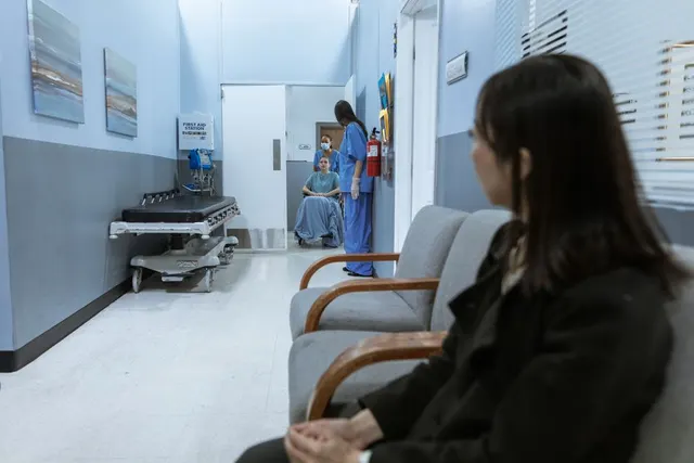 Femeie așezată într-un hol de spital, cu un pat de transport și personal medical în fundal
