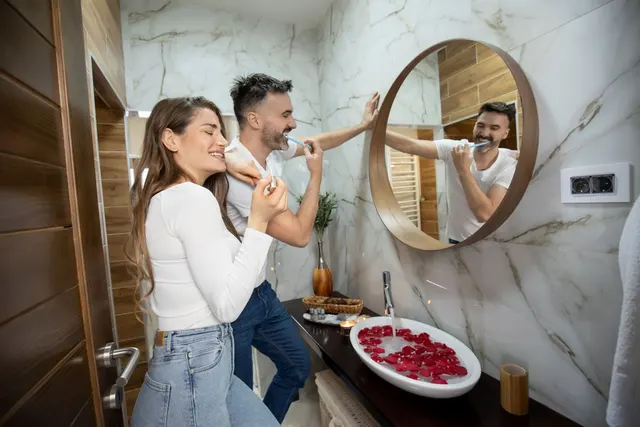 Cuplu tânăr râzând în fața oglinzii rotunde dintr-o baie cu pereți de marmură