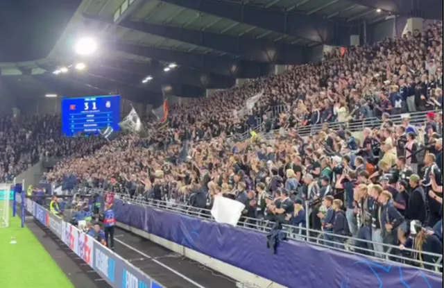 Tribune pline de suporteri pe un stadion, noaptea, cu tabela de marcaj aprinsă