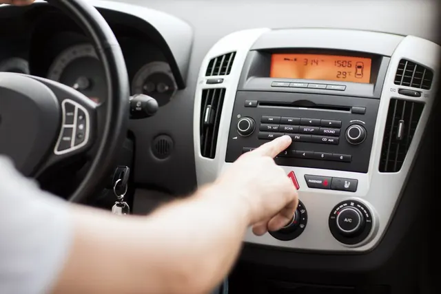 Mână care apasă un buton al radioului de pe bordul unei mașini, lângă volan