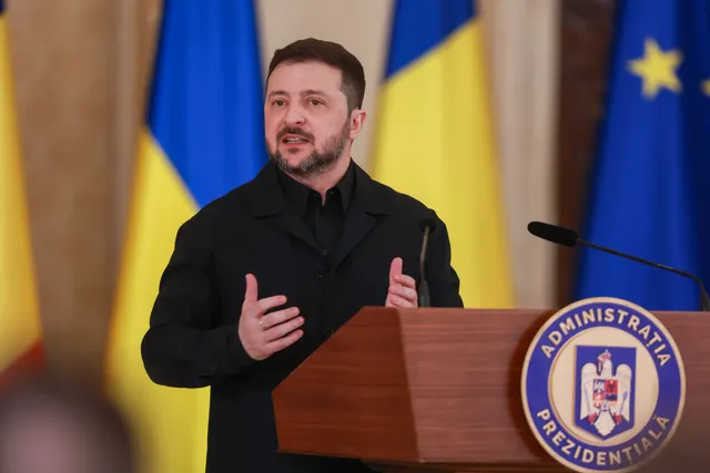 Bărbat cu barbă, în haine negre, vorbind la pupitrul Administrației Prezidențiale, între drapele