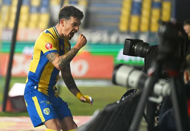 Fotbalist în echipament galben cu albastru sărbătorind cu pumnul strâns, lângă camere de filmat
