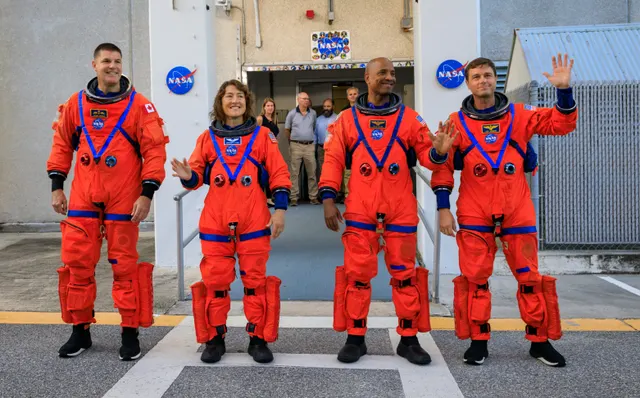 Patru astronauți în costume portocalii, salutând în fața unei clădiri cu sigla NASA