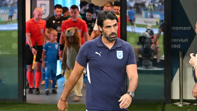 Bărbat în tricou polo bleumarin ieșind din tunelul stadionului, urmat de jucători și o mascotă