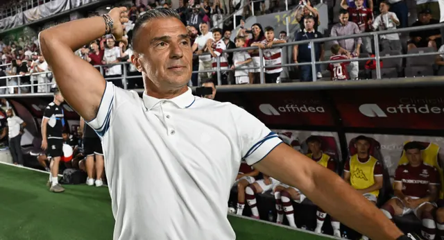 Bărbat în tricou polo alb, cu mâna la ceafă, lângă banca de rezerve a unui stadion