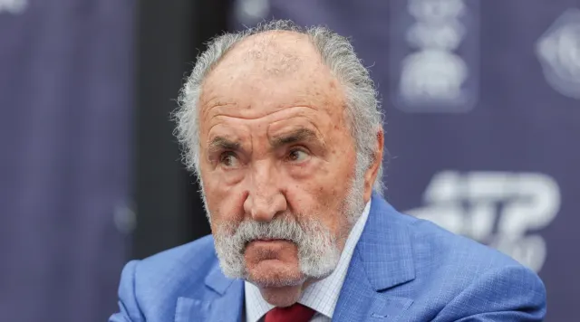 Bărbat în vârstă, cu părul alb și mustață, în sacou albastru, în fața unui panou ATP