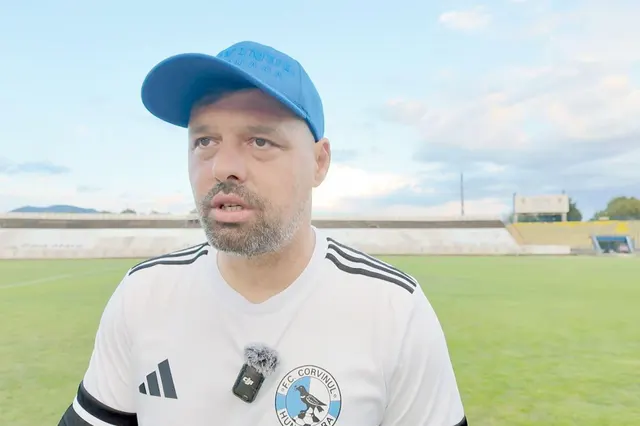 Bărbat cu șapcă albastră și tricou alb, cu microfon prins de guler, pe un stadion