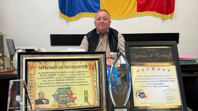 Bărbat în vârstă la biroul său, sub drapelul României, cu diplome de excelență pe masă