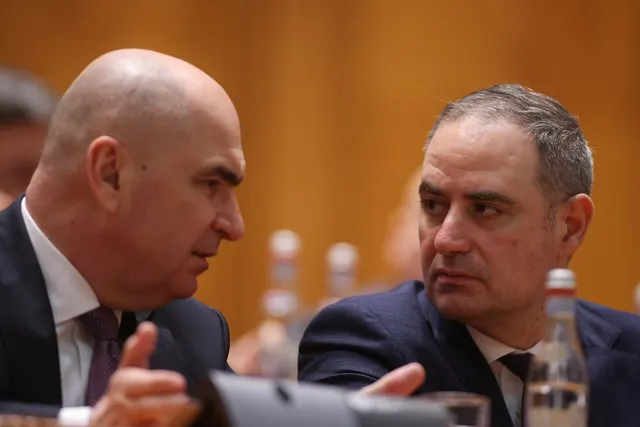 Doi bărbați în costume discutând la o masă de conferință, unul chel, aplecat spre celălalt