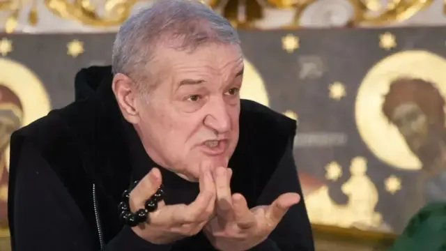Bărbat cărunt, în haină neagră, gesticulând cu ambele mâini în fața unor icoane aurite