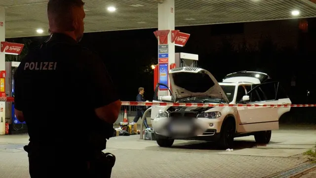 Polițist cu inscripția „Polizei” pe spate, lângă o mașină albă cu capota deschisă, la o benzinărie, noaptea