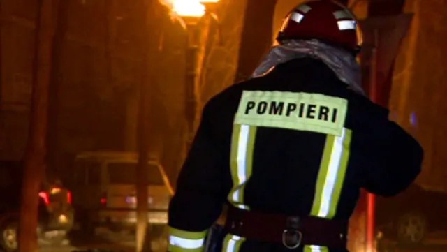 Pompier văzut din spate, cu inscripția „Pompieri” pe geacă, noaptea, lângă o clădire
