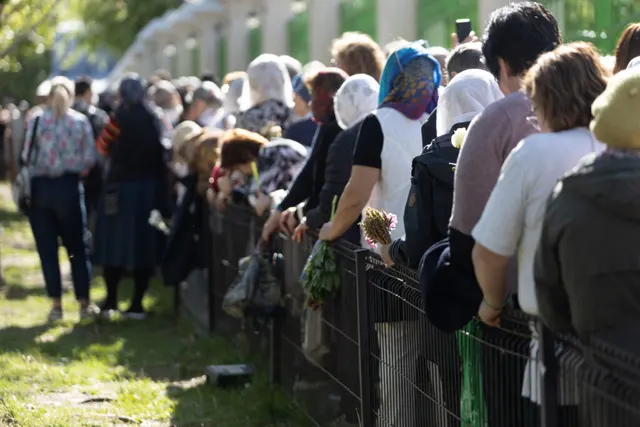Șir lung de oameni care așteaptă la coadă de-a lungul unui gard metalic, într-un parc