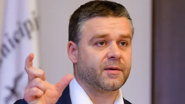 Bărbat în costum, cu barbă scurtă, gesticulând cu degetul ridicat în timp ce vorbește