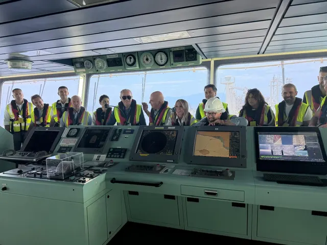 Grup de oameni în veste reflectorizante pe puntea de comandă a unei nave, în spatele pupitrelor de control