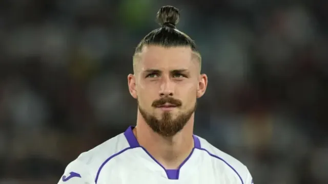Fotbalist cu părul prins în coc și barbă, în tricou alb cu guler violet