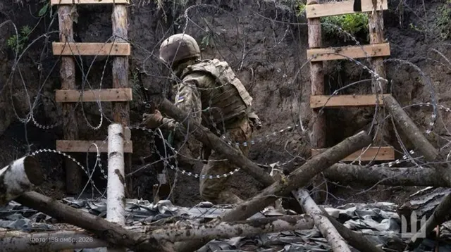 Militar în uniformă de camuflaj, ghemuit printre sârmă ghimpată și bușteni, într-o tranșee
