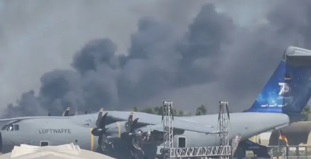 Captură de ecran: fum negru deasupra unui aerodrom, lângă un avion de transport cu inscripția „Luftwaffe”