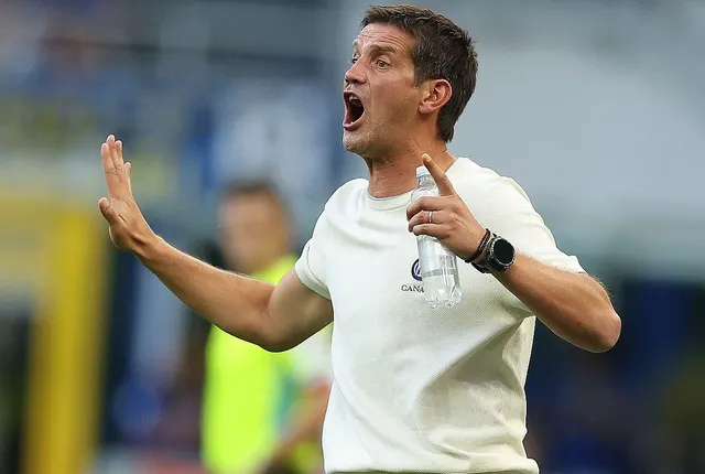 Bărbat în tricou alb gesticulând cu ambele mâini și strigând pe marginea unui teren de fotbal
