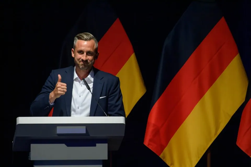 Bărbat în costum, cu degetul mare ridicat, la un pupitru, între drapelele Germaniei