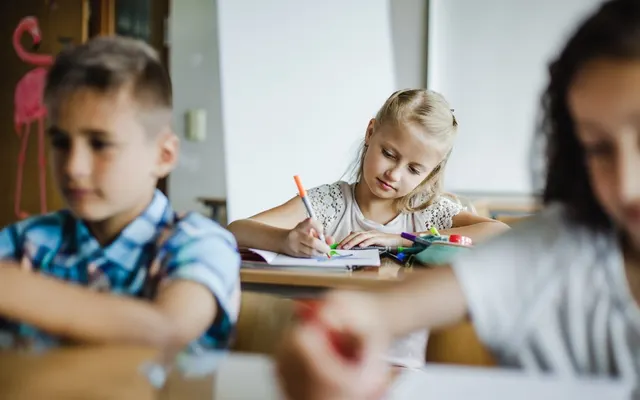 Copii într-o sală de clasă, desenând cu creioane colorate la bănci