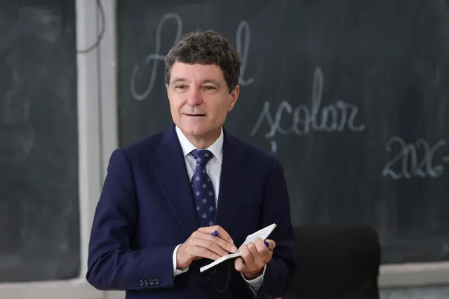 Bărbat în costum, cu un carnet în mână, în fața unei table pe care scrie cu creta despre anul școlar 2026
