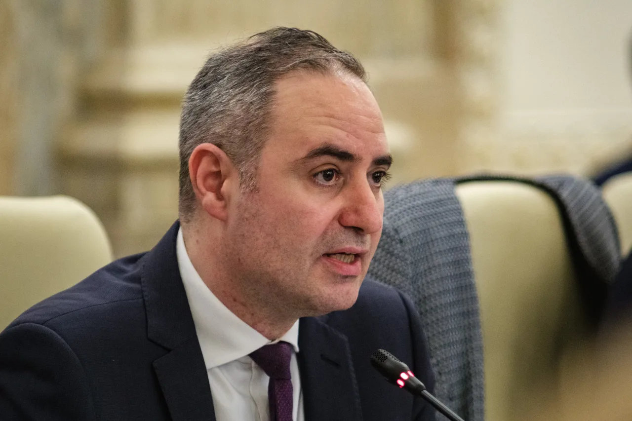 Bărbat în costum închis și cravată vișinie, vorbind la un microfon de masă