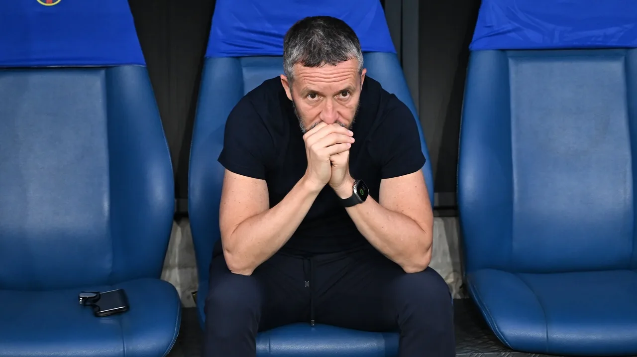 Bărbat în tricou negru, așezat pe un scaun albastru de stadion, cu mâinile împreunate la gură